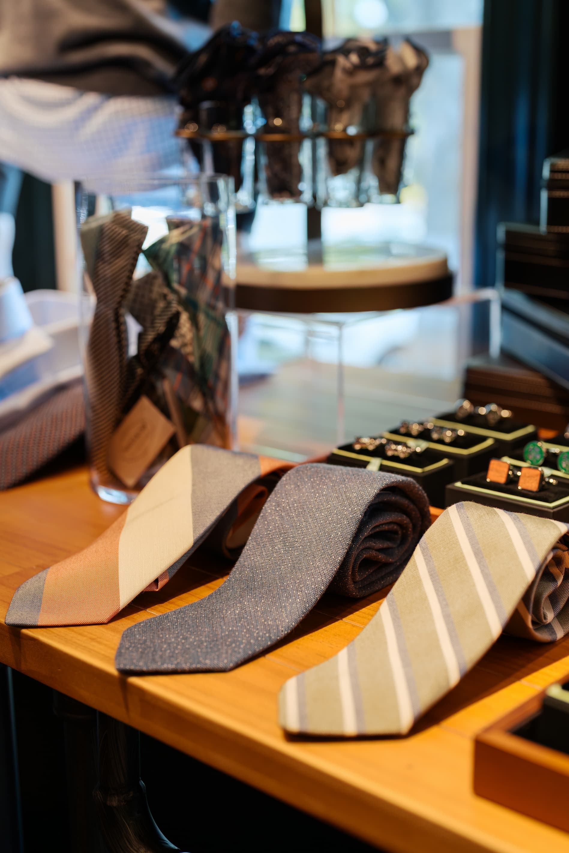 Close-up of a men’s boutique tabletop display featuring neatly rolled and laid neckties in soft, neutral patterns, with cufflinks and folded accessories arranged in the background.