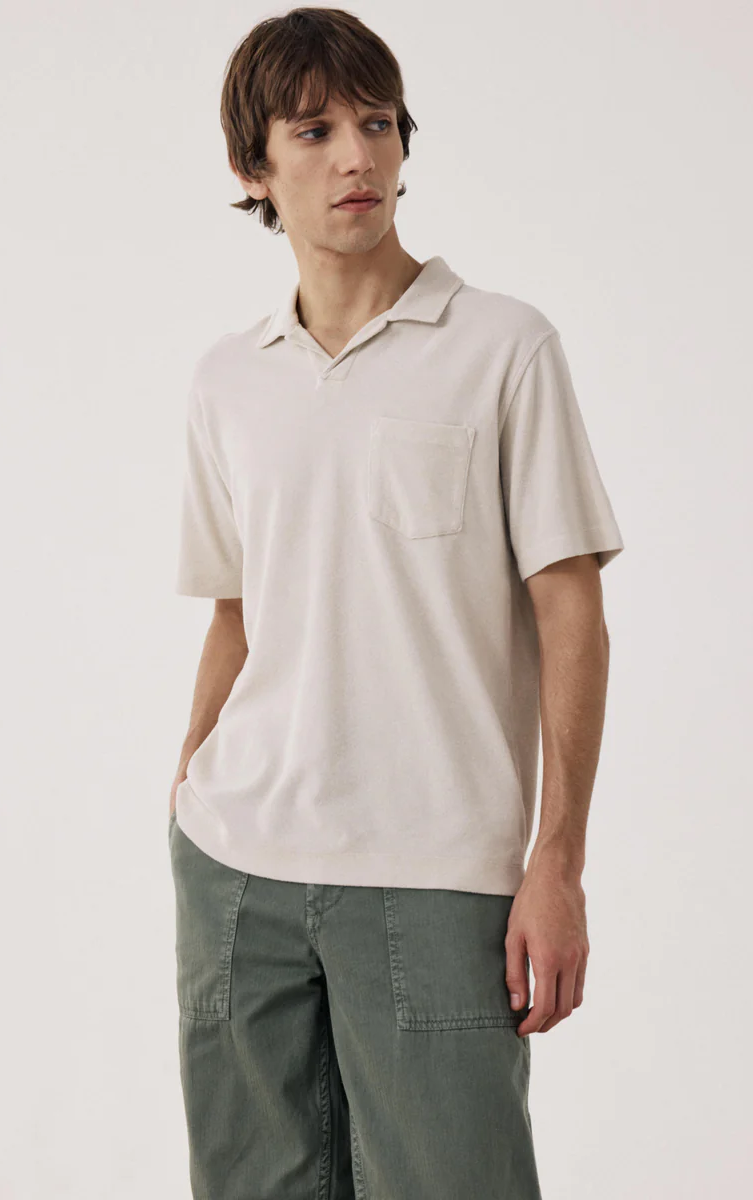 Man in a soft, neutral short-sleeve polo with a relaxed collar and chest pocket, paired with olive pants for an easy, understated look.
