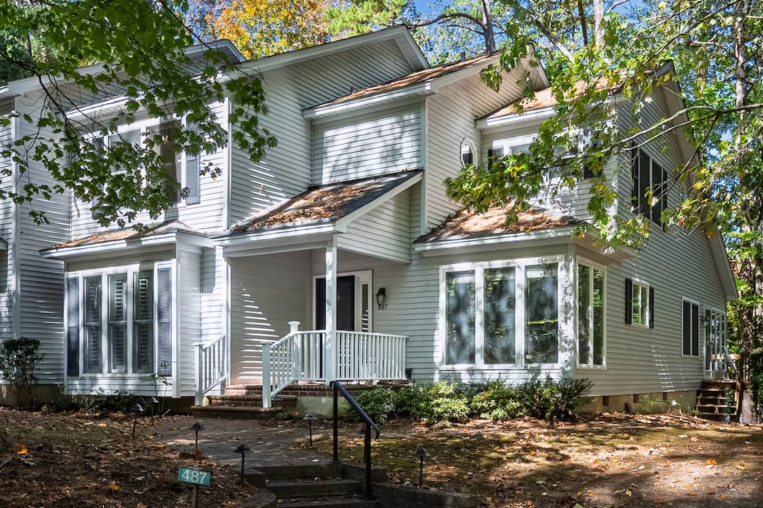 Two-story light gray clapboard townhome with a small covered porch, white railings, and a bay window, set beneath leafy trees along a shaded walkway.
