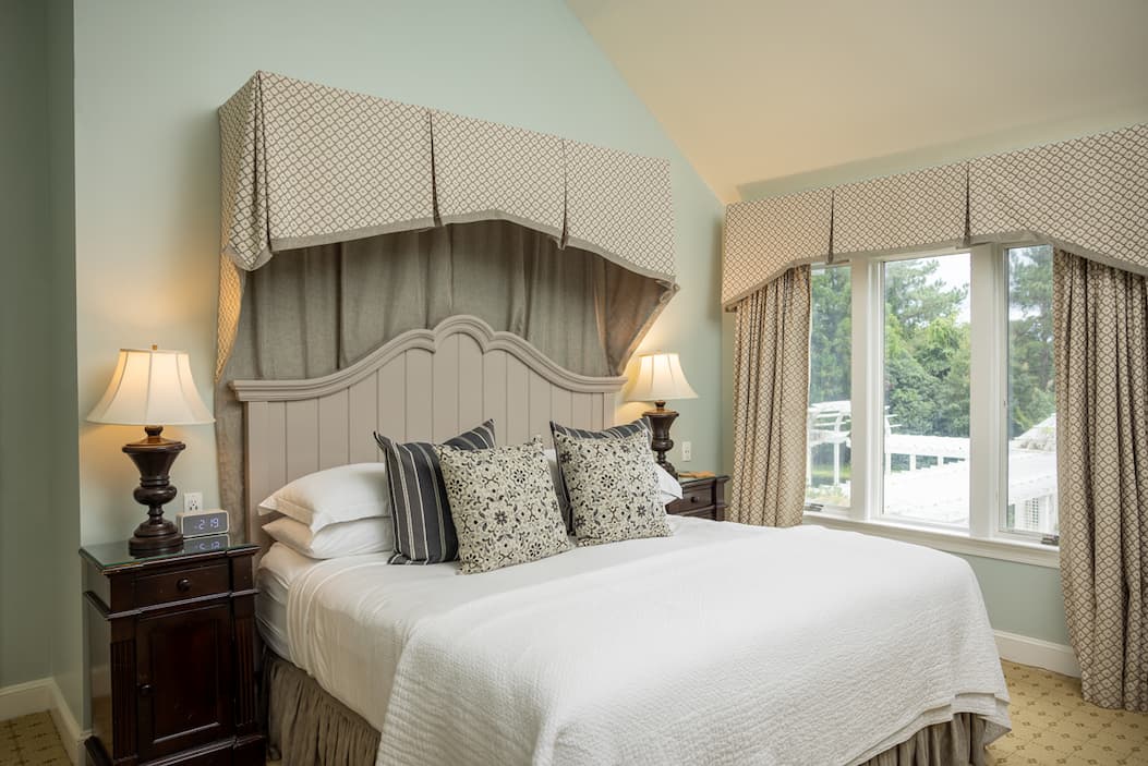 An elegant guest room features a king-size bed dressed in crisp white linens and layered patterned pillows beneath a decorative upholstered canopy. Soft sage-green walls, tailored drapery, and large windows create a bright, airy atmosphere with views of the surrounding grounds. Dark wood nightstands and classic table lamps add warmth and sophistication, while the traditional furnishings and refined details evoke a comfortable countryside retreat.