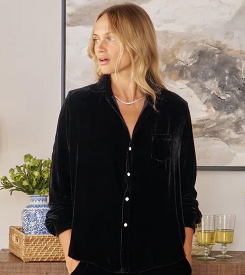 A woman wearing a black velvet button-up top stands indoors with her hands in her pockets, in front of a console table with decor including flowers, books, and glasses.