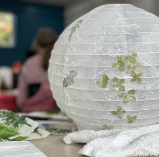 A white paper lantern decorated with green pressed leaves sits on a table, surrounded by craft supplies. In the blurred background, people are seated and working on similar projects in a classroom setting. Fearrington Village