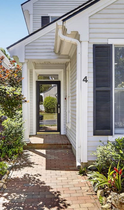 Brick walkway leading to a covered front entry with a black-framed glass door, white siding with black shutters, and neatly landscaped garden beds with shrubs and flowering plants on either side.