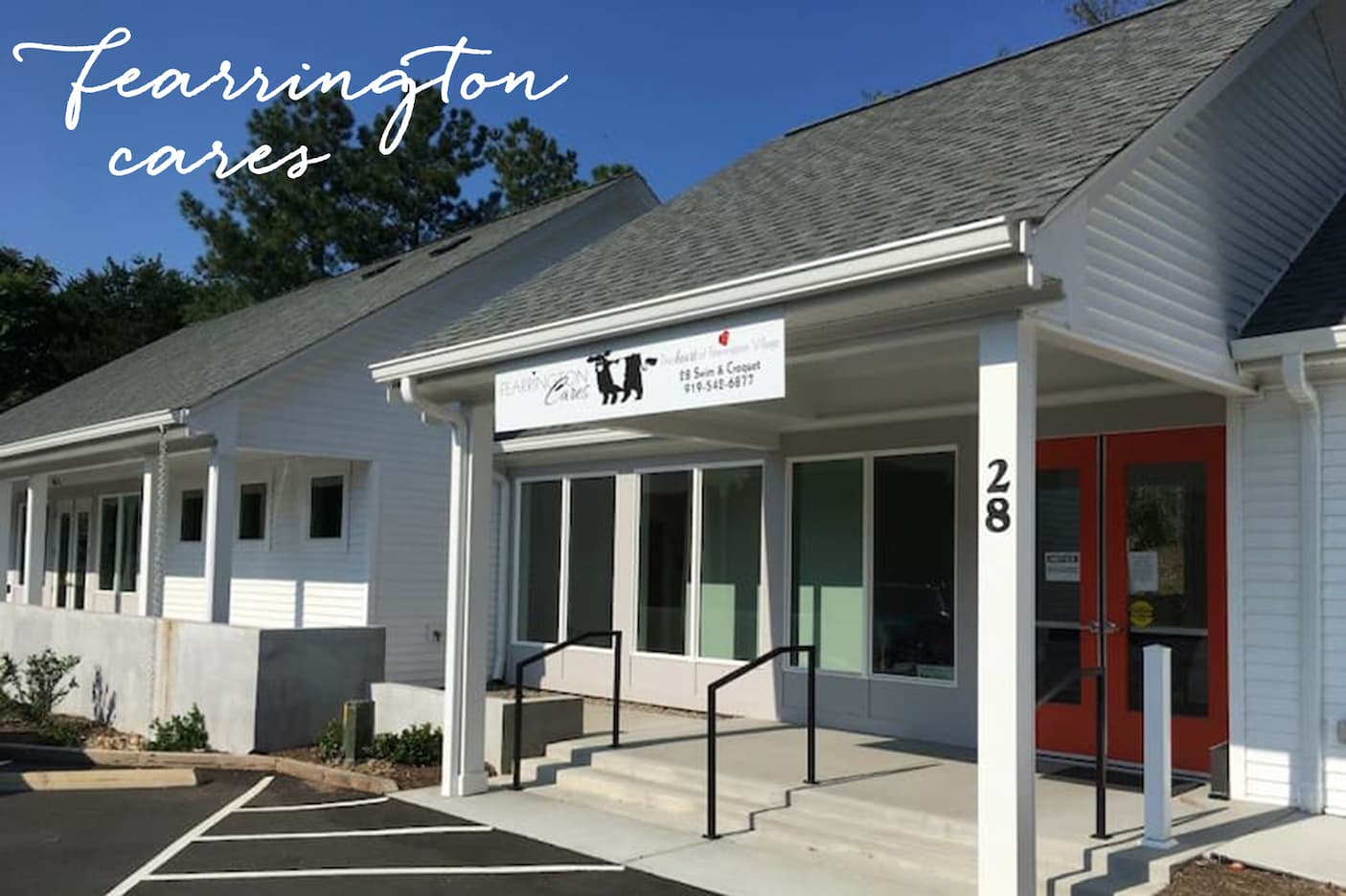Exterior of a white commercial building at Fearrington Village with a covered entry, steps and railings, red double doors, and signage for Fearrington Cares, photographed on a clear day.