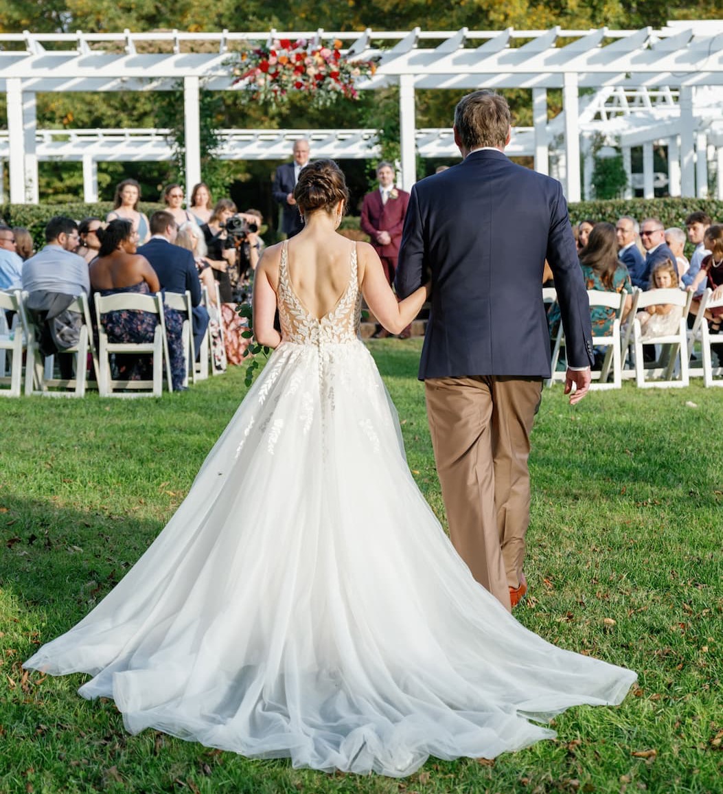 Bride in a flowing white gown walks arm in arm with an older man down a grassy aisle toward an outdoor wedding ceremony, surrounded by seated guests, white pergolas, and floral arrangements in a garden setting.