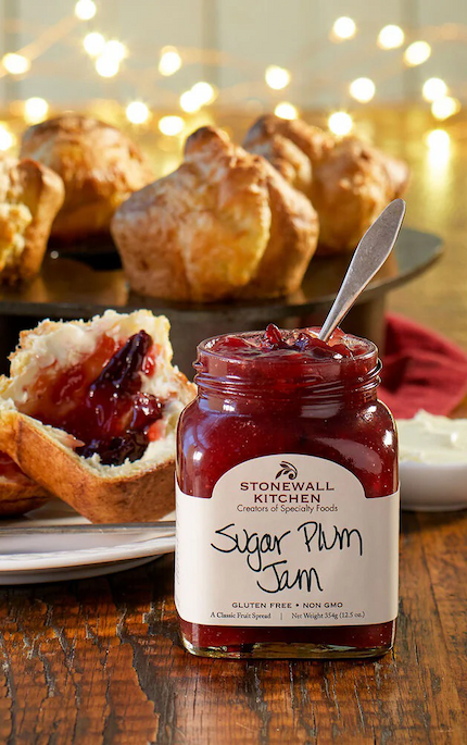 Jar of Stonewall Kitchen Sugar Plum Jam with a spoon inside, set on a wooden table beside a biscuit topped with jam, with golden pastries and soft holiday lights glowing in the background.
