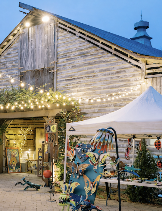 Rustic barn-style building lit with warm string lights at dusk, with an outdoor art market setup in front, including a white canopy tent, colorful metal sculptures, and a few people browsing in a cozy, festive courtyard.