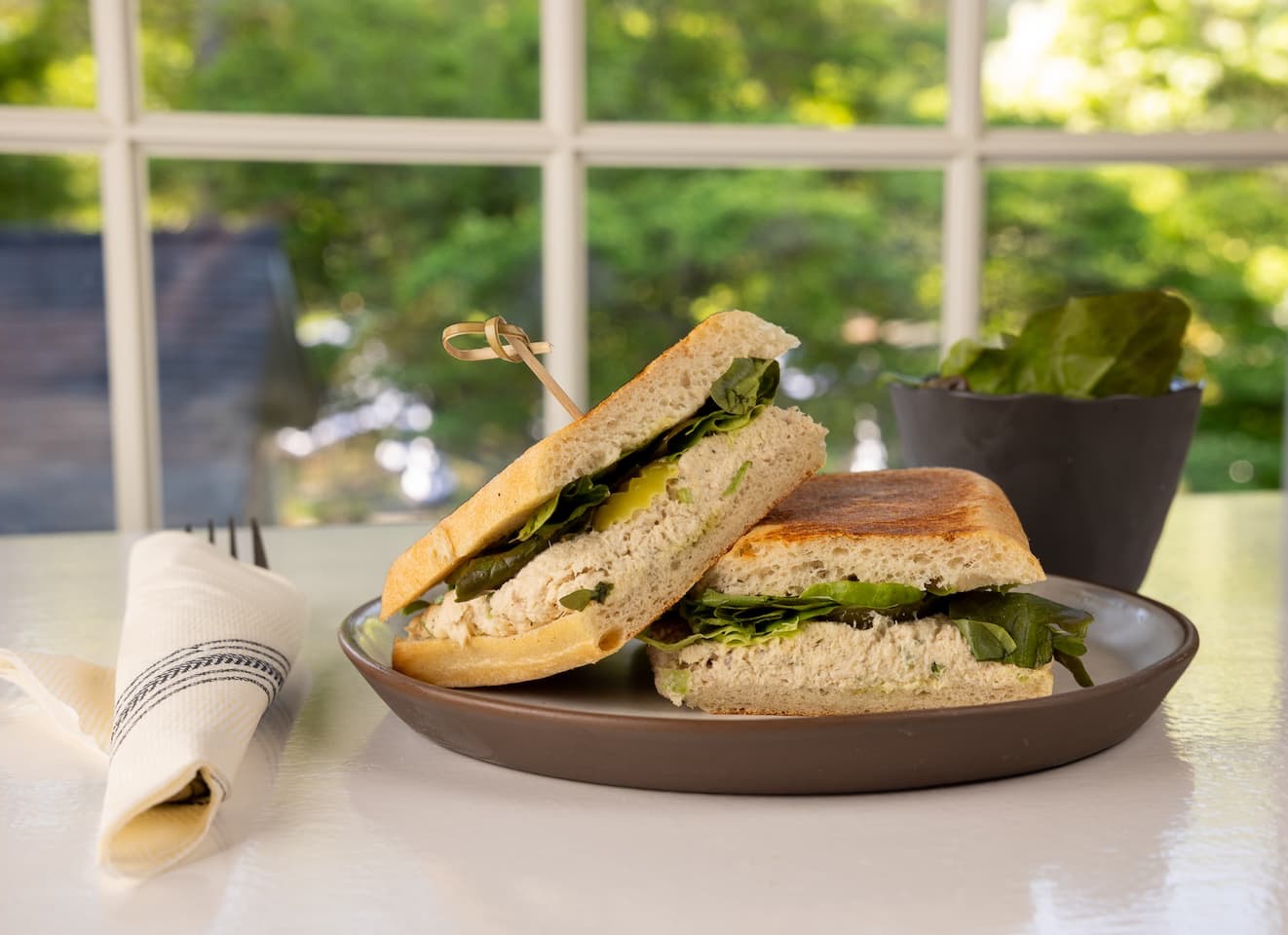A toasted sandwich filled with a creamy chicken salad and fresh greens is cut in half and stacked on a plate, secured with a skewer. It’s set beside a neatly rolled napkin and a small bowl of greens, with a bright window and leafy outdoor view in the background.