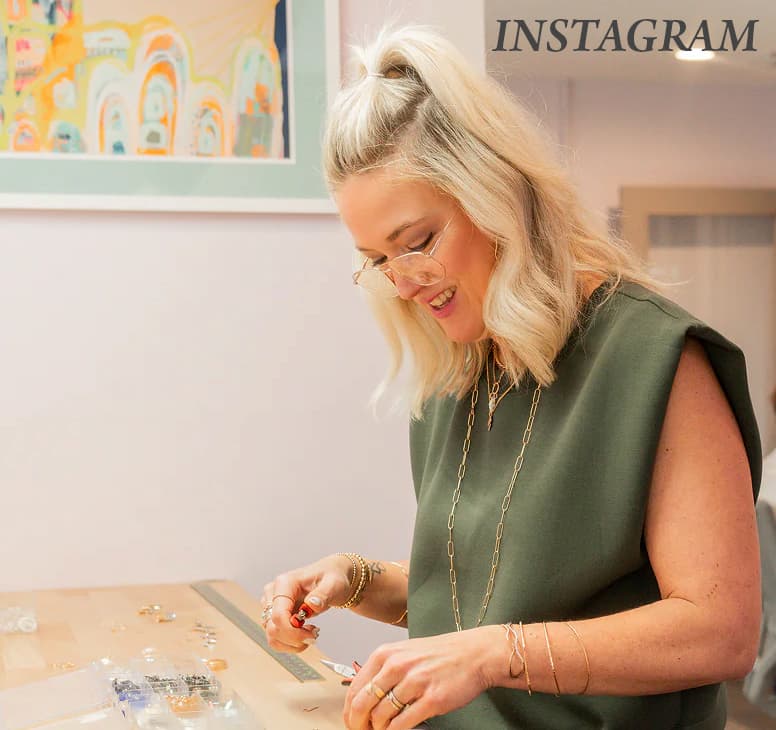 A person with light hair and glasses smiles while working at a jewelry-making table, arranging pieces and tools, with a sign reading “INSTAGRAM” visible on the wall behind them.