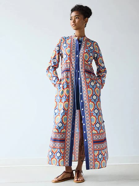 Full-length portrait of a woman wearing a vibrant patterned maxi shirt dress in blue, orange, red, and cream geometric prints. She stands barefoot in flat sandals against a soft neutral background, with hands in pockets and hair styled in a natural updo for an effortless, relaxed look.