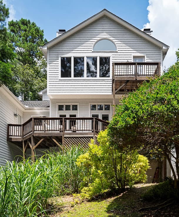 A wo-story house with gray siding and multiple wooden decks overlooking a landscaped backyard with shrubs and tall grasses.