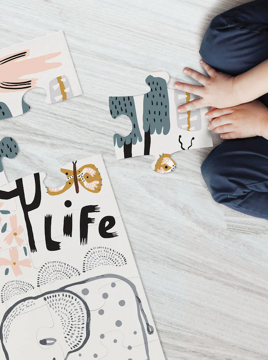 A child sitting on the floor works on assembling a colorful animal-themed puzzle featuring illustrations and the word "LIFE" in bold letters. Only the child's hands and legs are visible. Fearrington Village