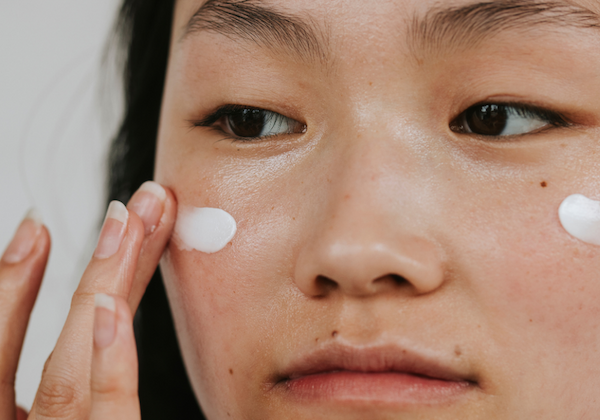 Close-up of a woman applying white facial cream to her cheeks with her fingertips. The image highlights smooth, hydrated skin and a skincare routine focused on moisturizing and nourishing the complexion.