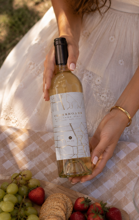 Top-down view of a woman in a white dress gently holding a bottle of Crossroads Sauvignon Blanc over a checkered picnic table, with grapes, strawberries, and crackers arranged nearby in soft natural light.