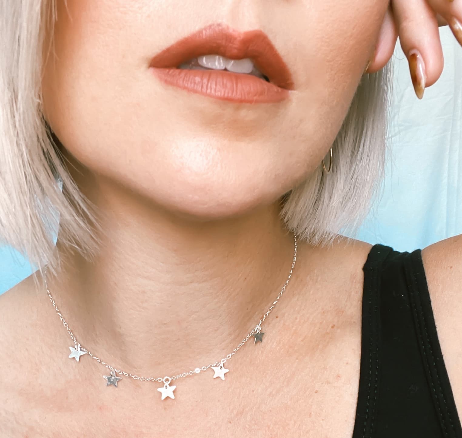 A close-up of a person’s lower face and neck wearing a delicate silver necklace with small star-shaped charms, paired with soft peach-toned lipstick and short, light-colored hair.