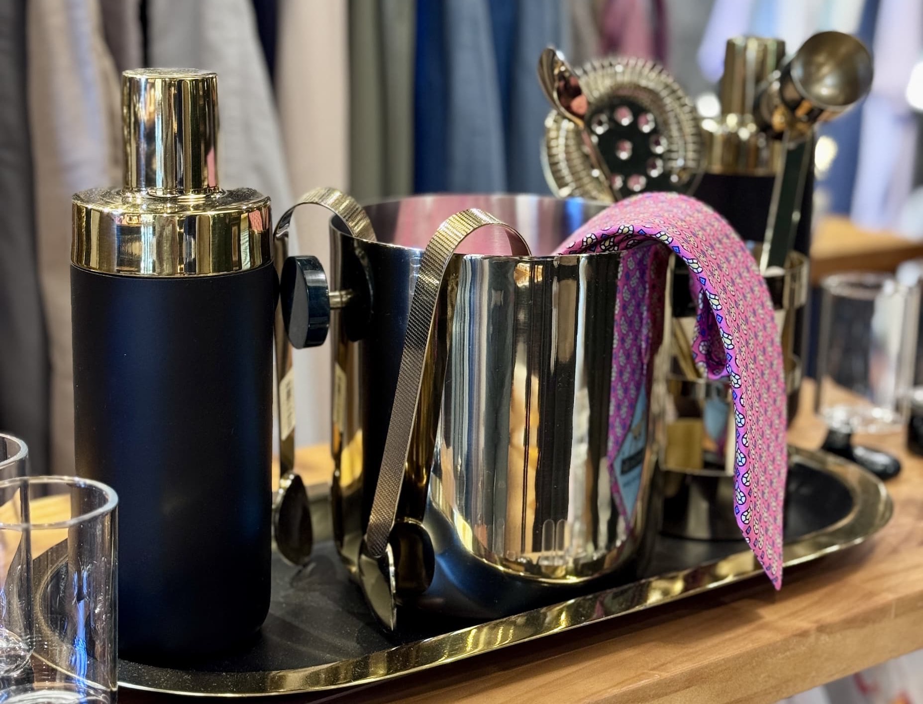Cocktail shaker, ice bucket, bar tools, and glassware displayed on a tabletop inside a home and gift shop with clothing in the background. Fearrington Village.