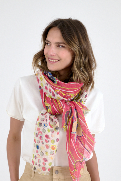 Woman wearing a white top and a colorful patterned scarf with pink stripes and multicolored dots looks to the side against a light background.