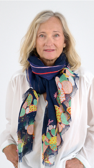 A woman with shoulder-length blonde hair wears a navy scarf with colorful embroidered designs over a white blouse, photographed against a simple light background.