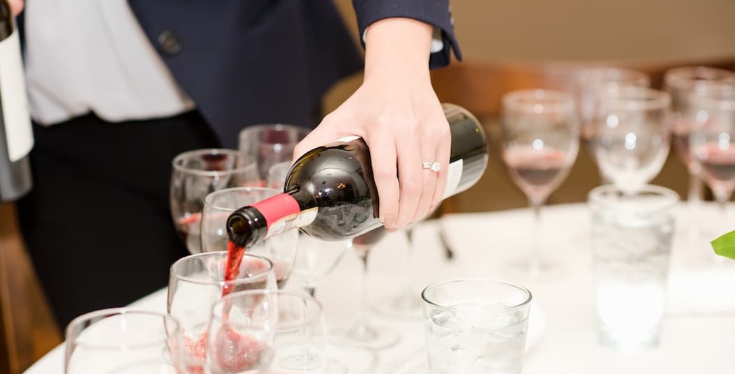 A wine tasting in progress as a server pours red wine from a bottle into a glass at a table set with multiple tasting glasses. Several partially filled glasses, a glass of ice water, and additional wine bottles create an inviting atmosphere for a guided tasting or wine-pairing event.