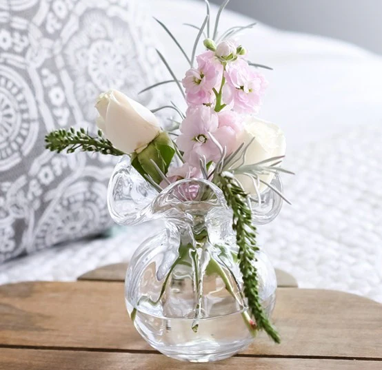 A small clear glass vase holds a delicate arrangement of pale roses and soft pink blossoms, set on a wooden table with a cozy, patterned backdrop.