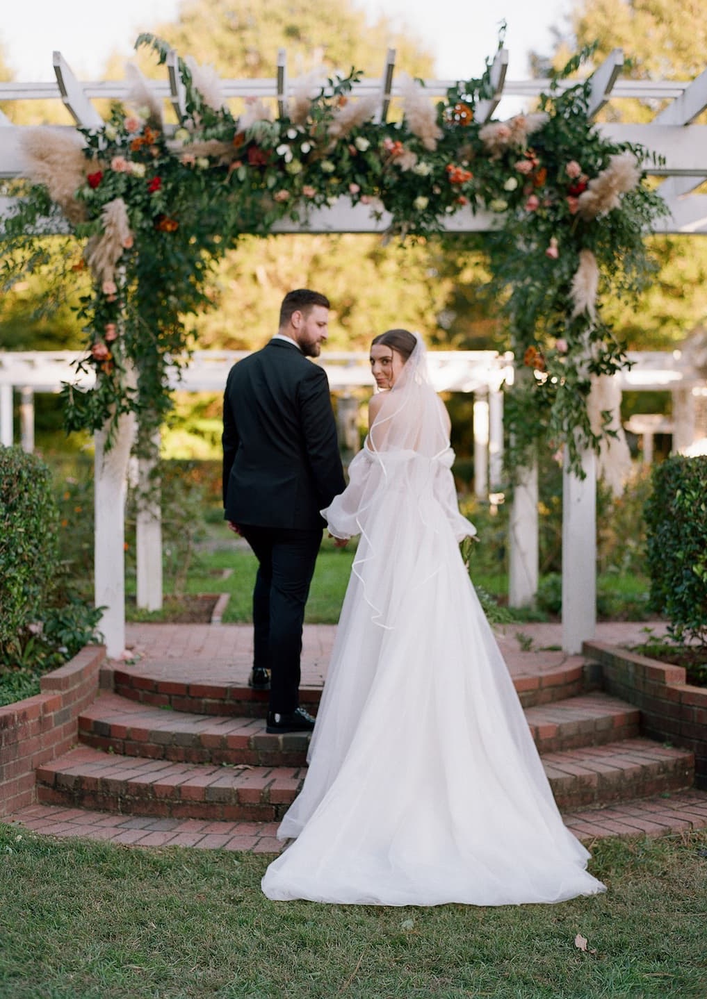 Bride in a flowing white gown and veil and groom in a dark suit walk beneath a flower-covered pergola in a garden, turning back toward the camera.