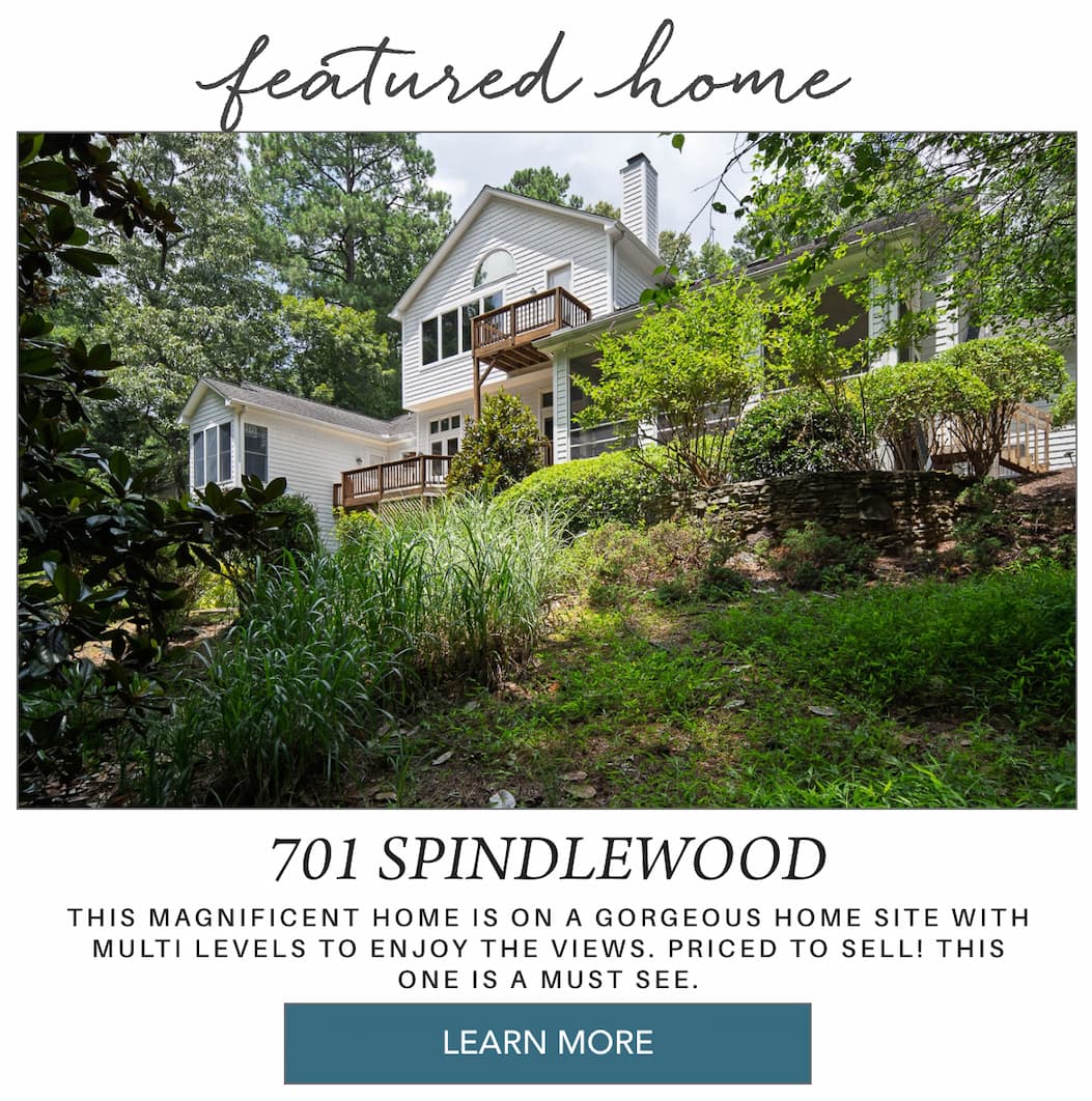 A two-story white house with a large wooden deck and balcony sits surrounded by trees and greenery on a sloped yard, advertised as a featured home for sale at 701 Spindlewood. Fearrington Village