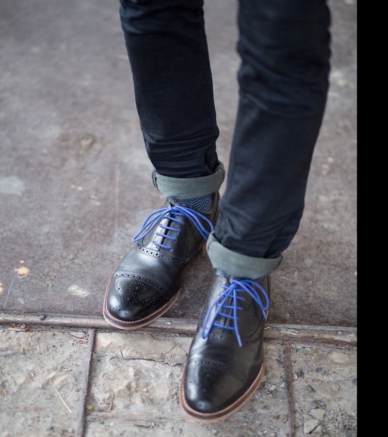 A person wearing black leather brogue shoes with bright blue laces, standing on a stone and concrete surface with jeans cuffed at the ankles.