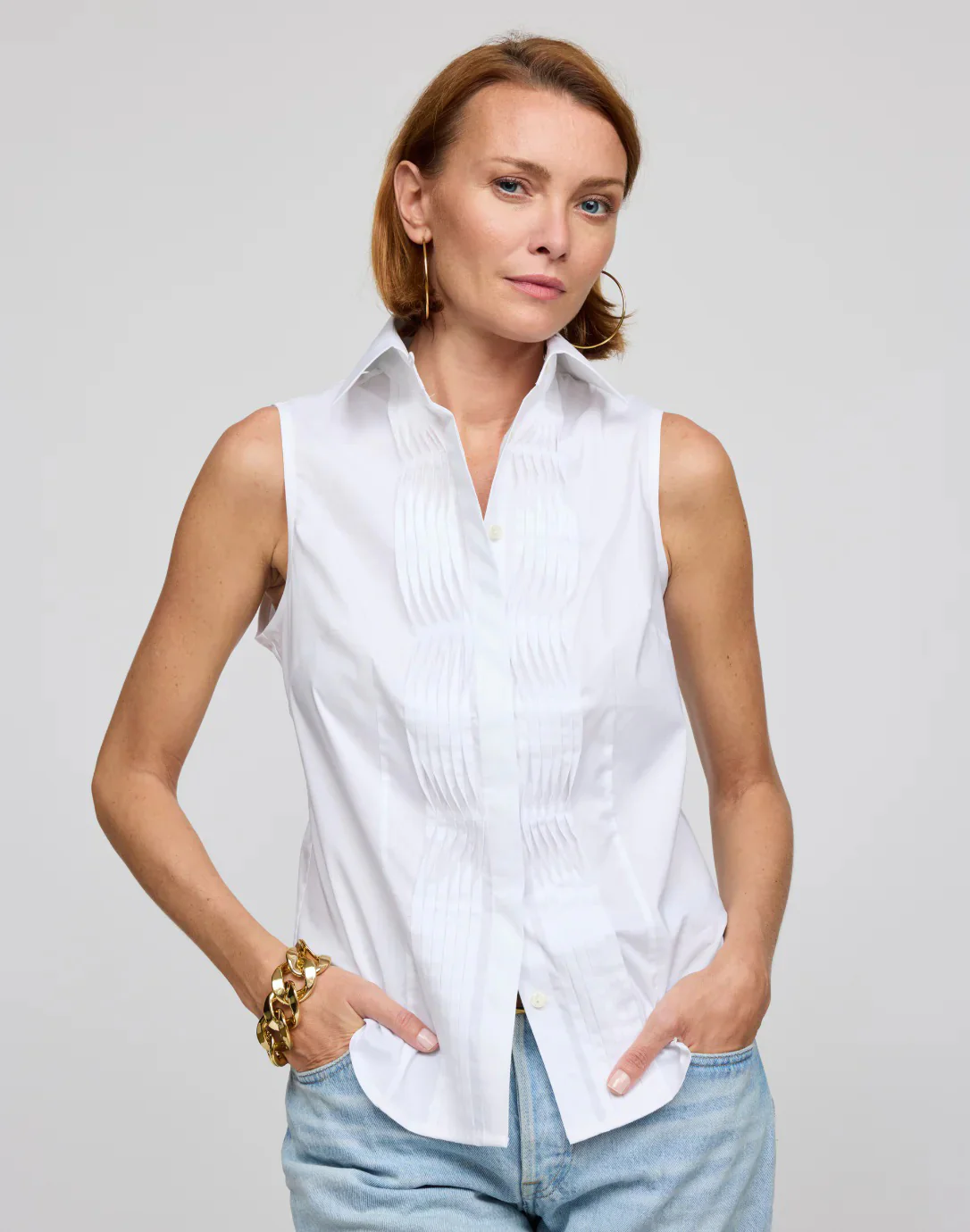 A woman stands against a light gray background wearing a sleeveless white button-front blouse with pleated detailing and light blue jeans. She has short auburn hair, gold hoop earrings, and a chunky gold bracelet, with her hands casually tucked into her pockets.