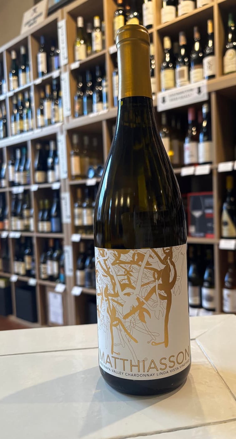 A bottle of Matthiasson Linda Vista Vineyard Chardonnay stands on a white-tiled counter in front of a wall of neatly arranged wine shelves. The elegant label features gold and cream botanical artwork, while the softly blurred background of wine bottles creates a warm, inviting wine shop atmosphere. The image highlights the bottle's distinctive design and premium presentation.
