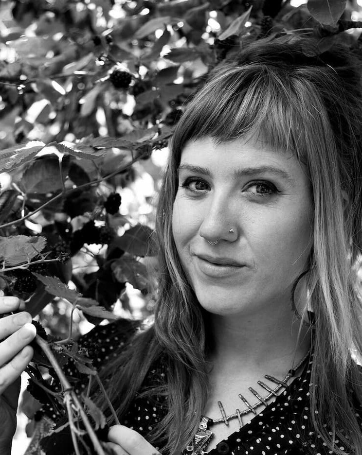 Black-and-white portrait of author Rebecca Beyer standing among leafy branches, holding a stem and looking toward the camera.