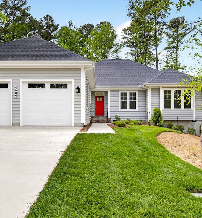 Single-story gray house with white trim, a red front door, attached garage, and neatly landscaped lawn bordered by trees.