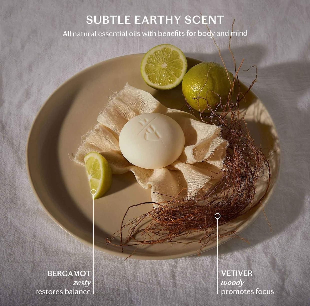 Round lotion bar on a plate with lime slices and vetiver roots, illustrating its subtle earthy scent from bergamot and vetiver essential oils.