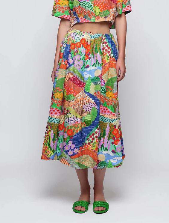 Woman wearing a colorful, patterned midi skirt with a matching cropped top, featuring a vibrant landscape-inspired print, paired with green sandals.