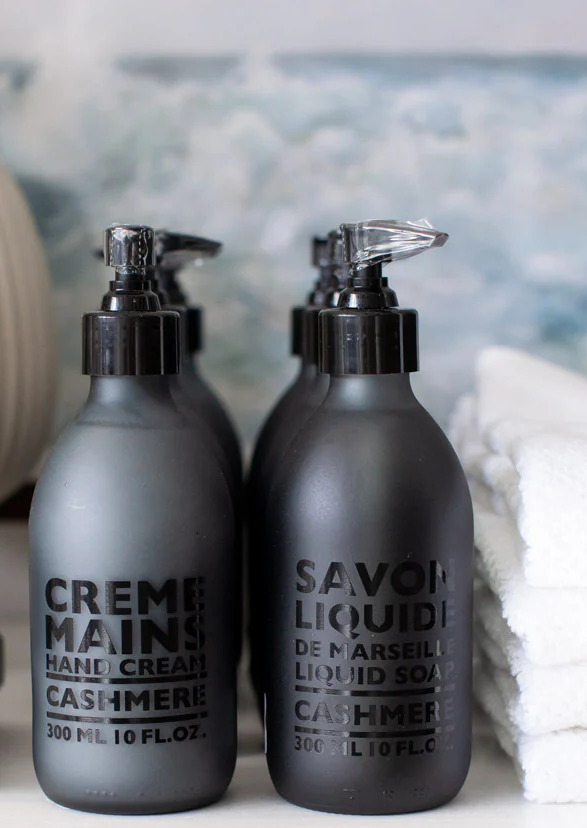 Two matte black pump bottles labeled hand cream and liquid soap, set beside neatly folded white towels on a bathroom counter.