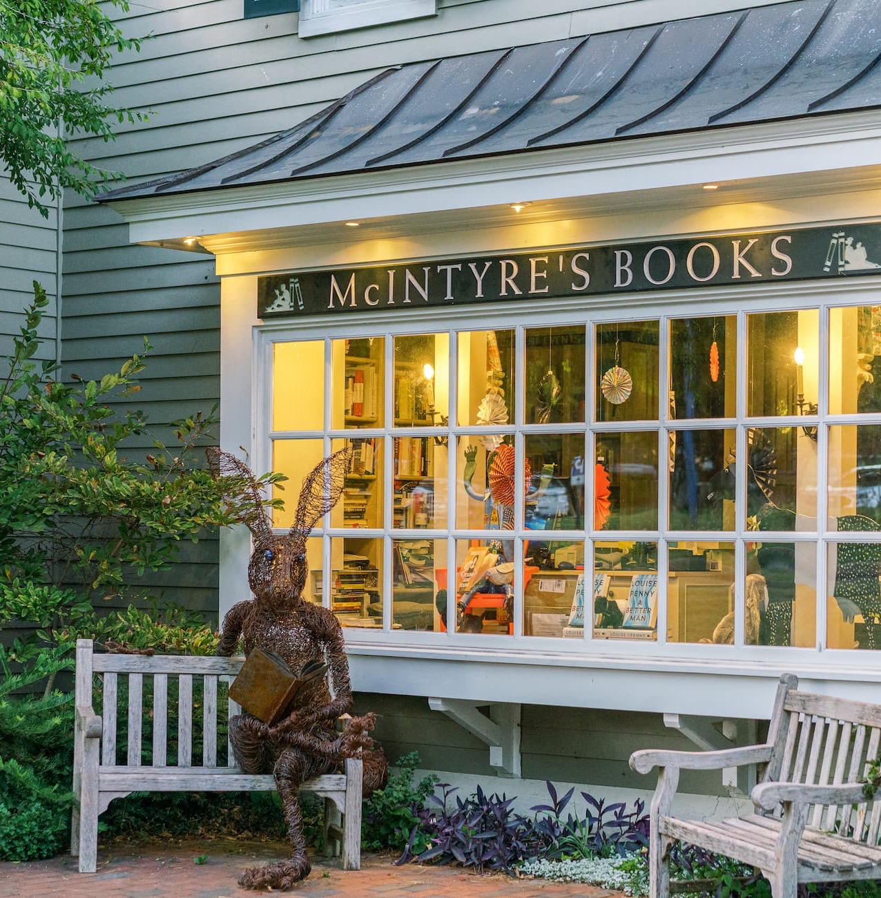 Exterior of McIntyre’s Books at Fearrington Village, with a whimsical metal rabbit sculpture reading on a bench outside the warmly lit shop windows.