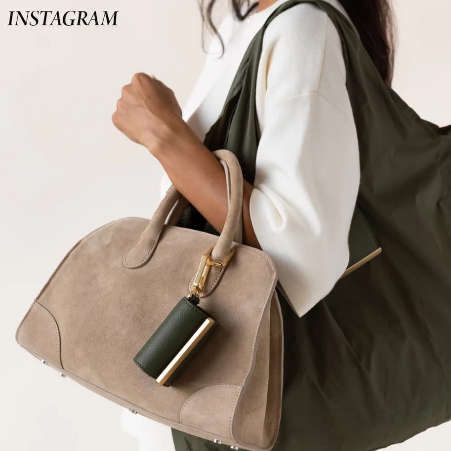 A woman carries a taupe suede handbag and an oversized olive-green tote bag, both accented with gold-tone hardware and a matching leather accessory pouch. Styled with a relaxed white top, the image showcases versatile, modern bags designed for everyday use and travel.