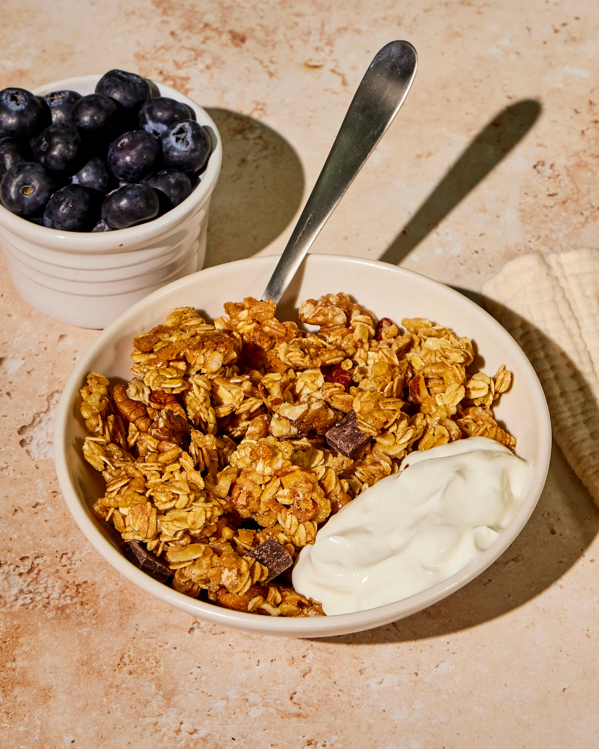 A bowl of golden oat granola with nuts and chocolate pieces sits on a stone surface, topped with a spoonful of yogurt, with a small ramekin of fresh blueberries beside it and a spoon resting in the bowl.