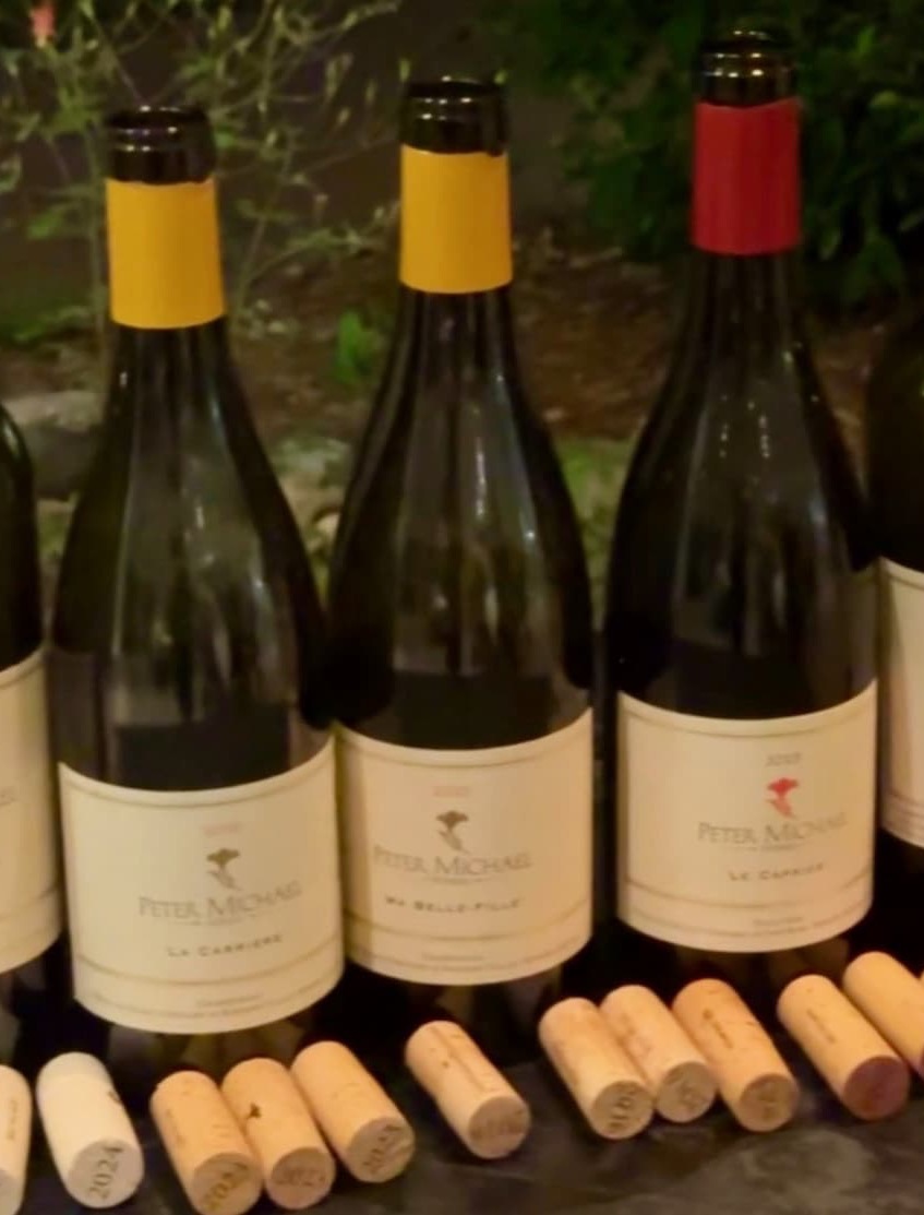 Three bottles of Peter Michael wine are displayed side by side on a tasting table, each distinguished by a different capsule color. Corks from opened bottles are arranged in front, while soft evening lighting and garden greenery in the background create an intimate wine tasting setting.