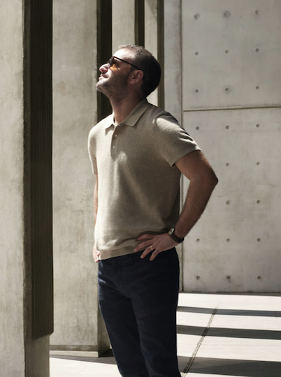 Man in a light beige polo and dark pants stands in a modern concrete space, looking upward with one hand on his hip, sunlight casting strong architectural shadows across the scene.