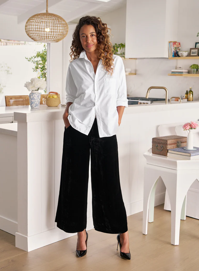 A woman stands in a bright kitchen wearing a white button-up shirt and black wide-leg pants, with her hands in her pockets and black heels on.