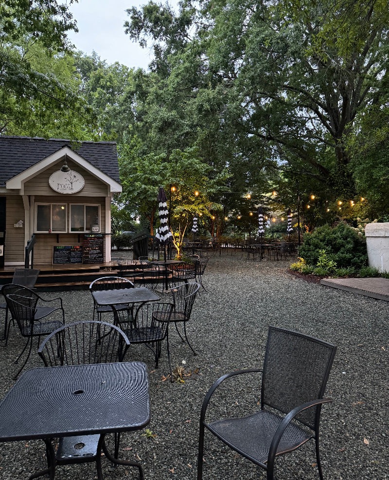 A quaint outdoor café with a small wooden building featuring a menu board and sign that reads "Food". The seating area has wrought-iron tables and chairs on a gravel ground. Black-and-white striped umbrellas are scattered around. Tall green trees surround the serene setting. Fearrington Village