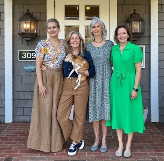 Four women stand on the brick steps of a yellow-sided house with a dark green front door. They are casually posed and smiling, dressed in fall-toned outfits. One woman on the right is slightly elevated on the porch and is holding a small dog in an orange sweater. The scene feels warm, friendly, and welcoming, like a relaxed front-porch portrait.