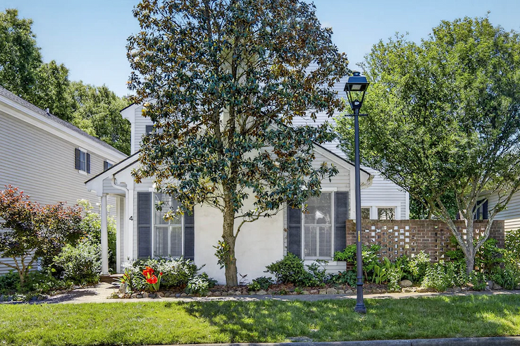 A charming cottage-style home sits behind a lush front garden filled with flowering plants, shrubs, and mature trees. A large magnolia tree anchors the landscape, partially framing the white exterior and gray shutters. A brick privacy wall, classic streetlamp, and neatly manicured lawn add to the neighborhood's inviting character, while bright sunshine and clear skies highlight the home's peaceful, well-kept setting.
