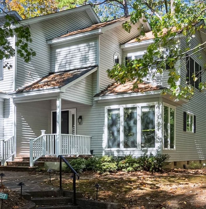 Two-story light gray clapboard townhome with a small covered porch, white railings, and a bay window, set beneath leafy trees along a shaded walkway.