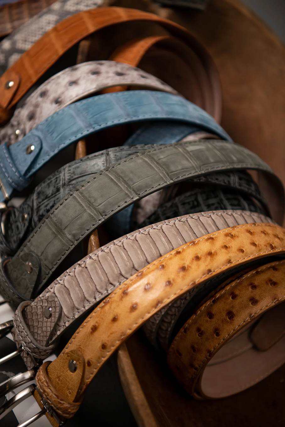 Assorted textured leather belts in shades of tan, gray, blue, and brown draped over a wooden stand, showing embossed patterns and stitched detailing.