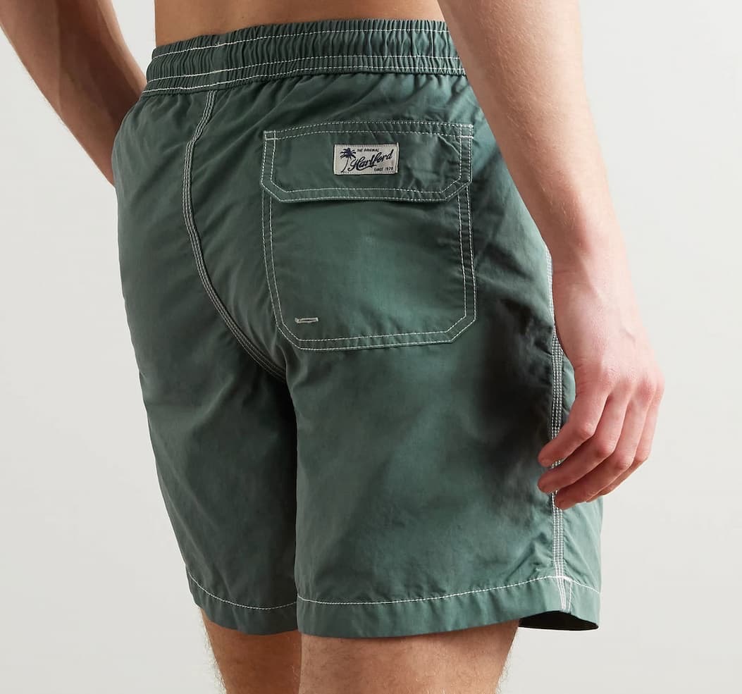 Men’s deep green swim trunks with contrast white stitching, elastic waistband, and back flap pocket detail, shown from the back against a light gray background.