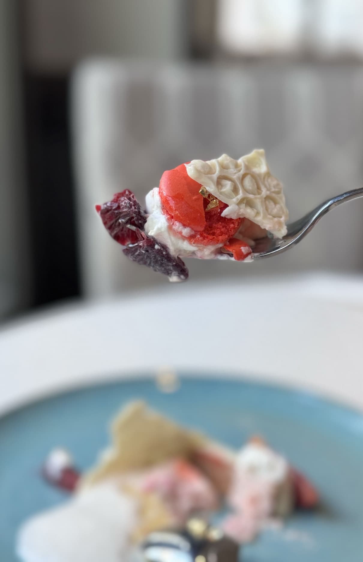 Close-up of a spoon holding a delicate dessert bite with red sorbet, whipped cream, candied fruit, and a crisp white garnish, with the plated dessert blurred in the background.