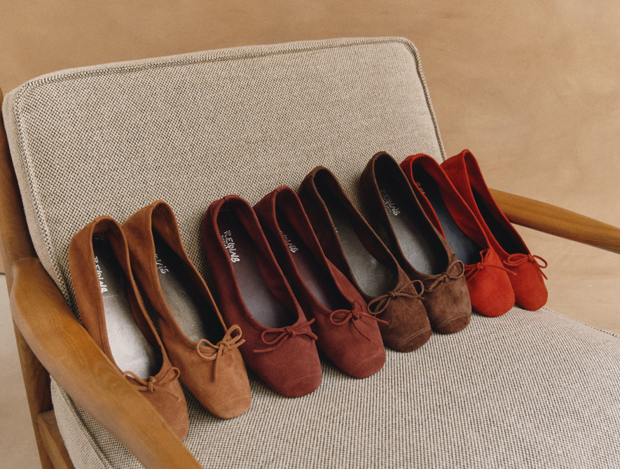 Five pairs of suede ballet flats in shades of brown and red are neatly lined up on a beige, mid-century modern armchair with wooden armrests, against a matching brown background. Fearrington Village