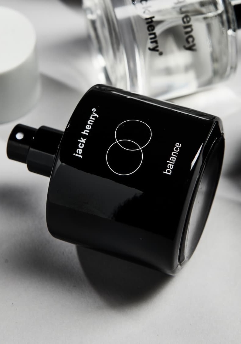 Black Jack Henry “Balance” fragrance bottle lying on its side beside a clear glass bottle on a light gray surface.