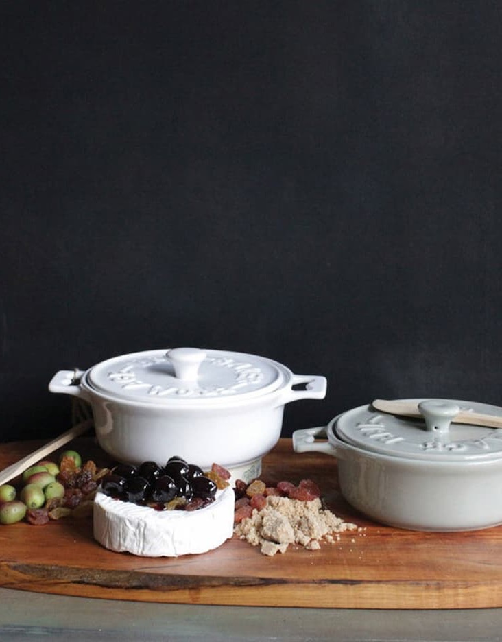 Two ceramic brie bakers with fitted lids rest on a rustic wooden serving board alongside baked brie topped with dark cherries, olives, dried fruit, and crumbs. A wooden spreader and moody dark backdrop create a warm, elegant entertaining scene.